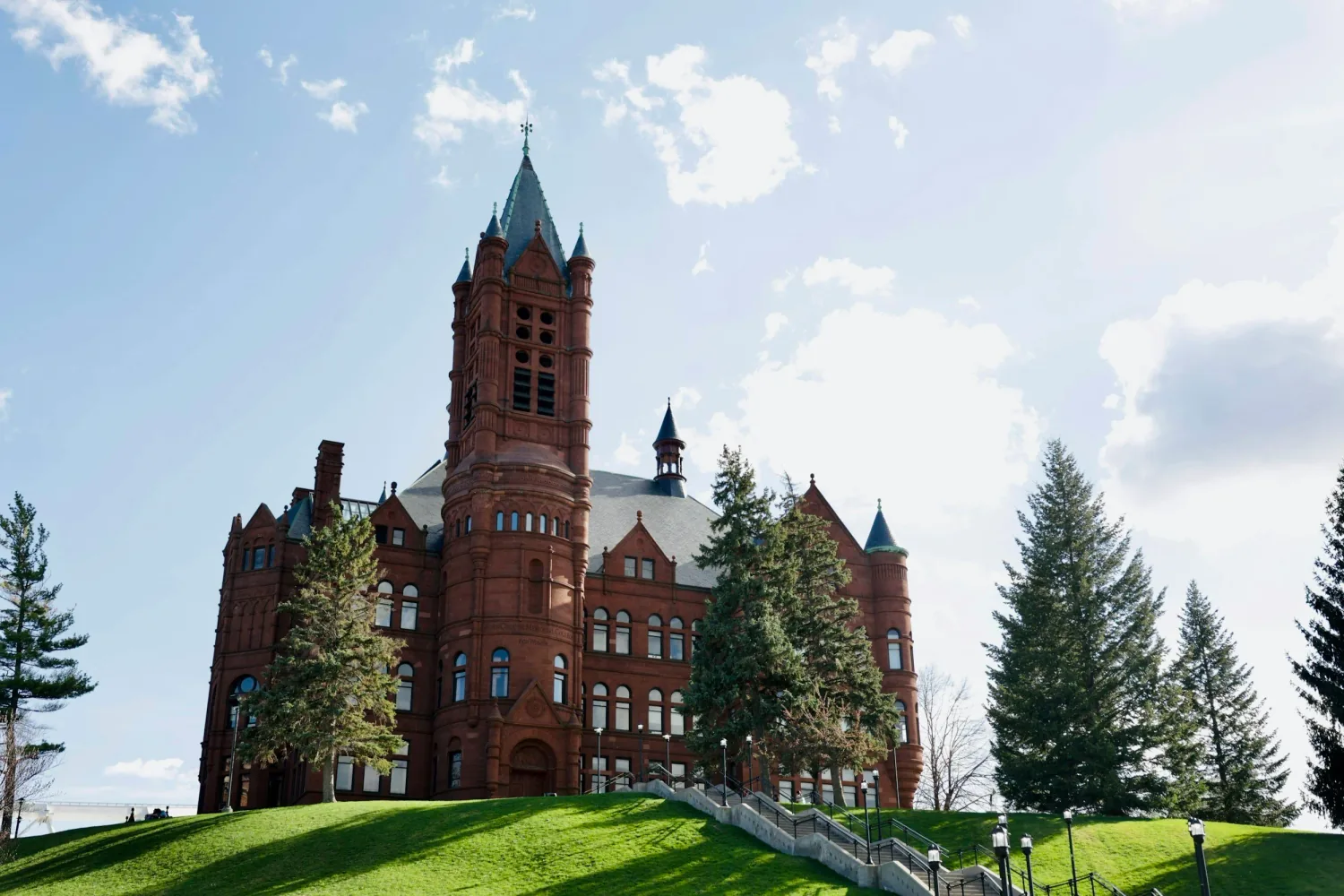 A large red brick castle with towers sits atop a hill with a green lawn and trees, under a partly cloudy blue sky.