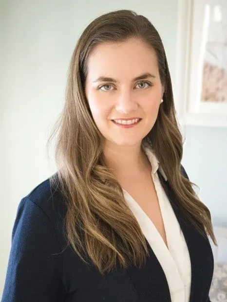 A young woman with long, wavy brown hair, wearing a navy blazer and white blouse, smiling in an indoor setting with a window in the background.